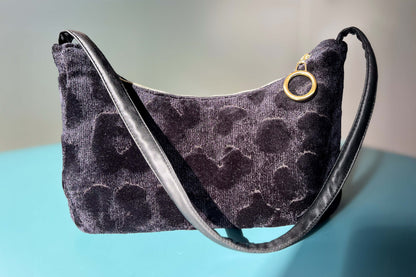 Gesamtansicht: Schultertasche im Vintagestil vor blau-grauem Hintergrund. Die Tasche ist schwarz gemustert und aus Samt und hat einen goldenen Reißverschluss. Die Tasche wurde aus upcycled Materialien von Jeff Every Piece A Friend hergestellt