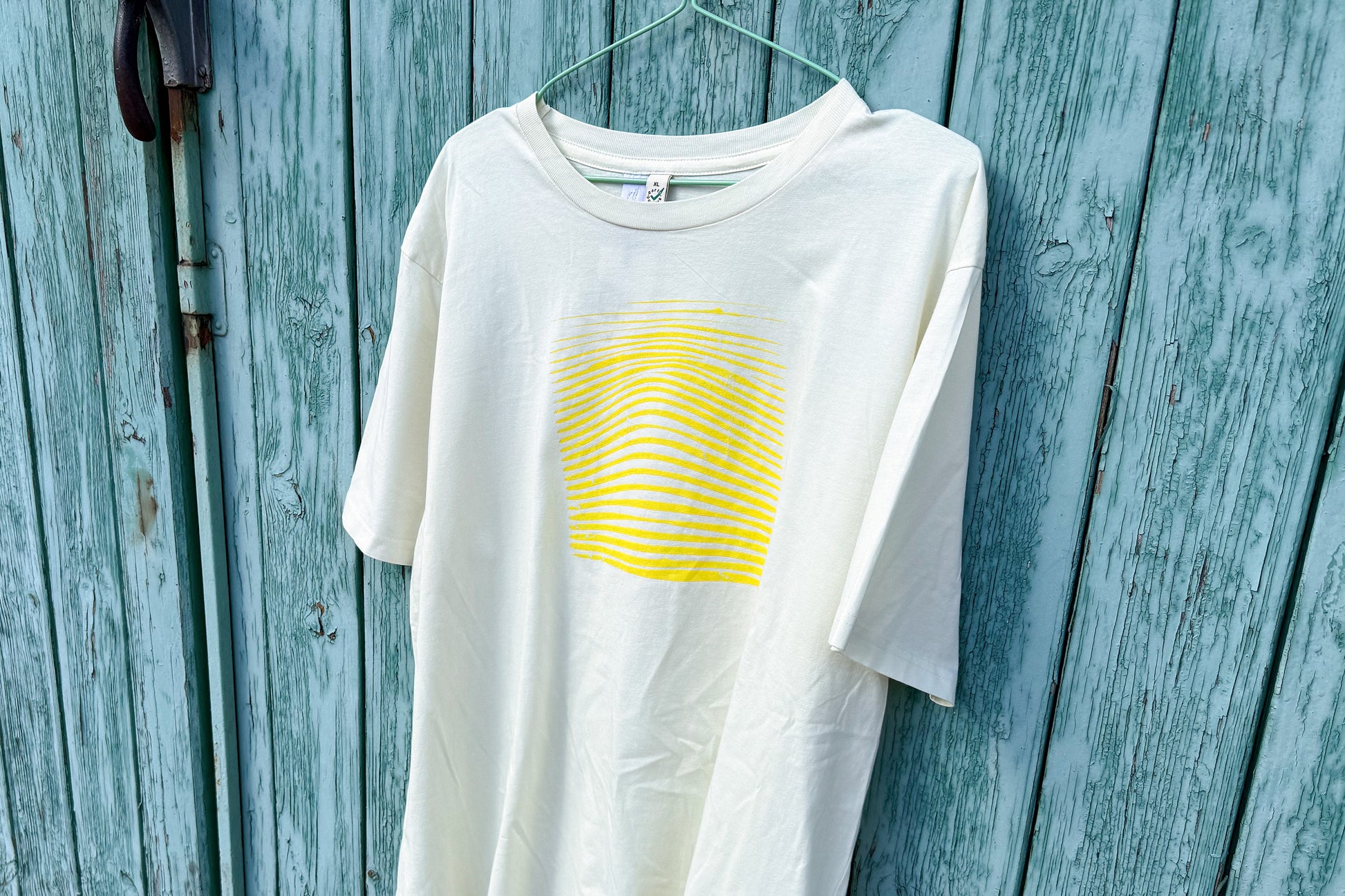 Detailansicht von einem hellgelben T-Shirt vor in der Größe XL vor einem blauen Hintergrund. Das T-Shirt hat einen welligen, neongelben Druck auf der Brust. Handbedruckt von Jeff - EveryPieceAFriend