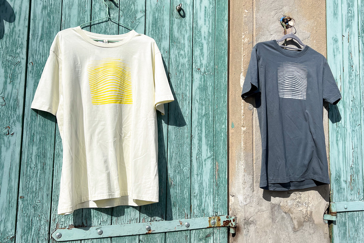 Ansicht von einem grauen und einem hellgelben T-Shirt vor einem blau-beigen Hintergrund. Das graue T-Shirt hat einen welligen Druck in silber, das hellgelbe Shirt einen welligen Druck in gelb auf der Brust. Handbedruckt von Jeff - EveryPieceAFriend