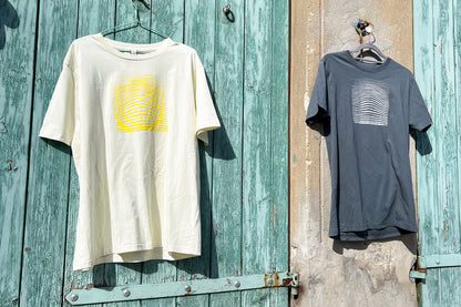 Ansicht von einem grauen und einem hellgelben T-Shirt vor einem blau-beigen Hintergrund. Das graue T-Shirt hat einen welligen Druck in silber, das hellgelbe Shirt einen welligen Druck in gelb auf der Brust. Handbedruckt von Jeff - EveryPieceAFriend