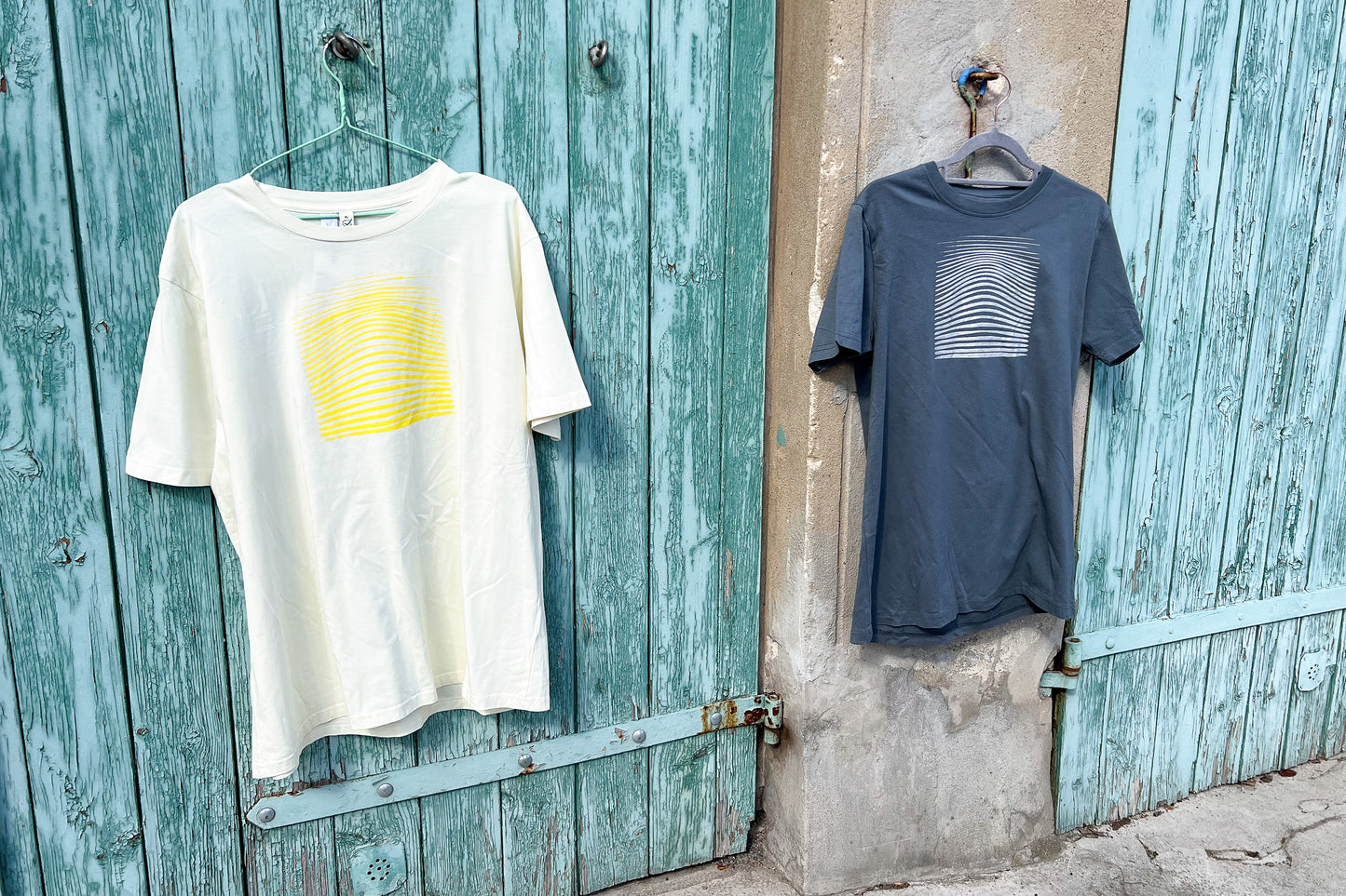 Ansicht von einem grauen und einem hellgelben T-Shirt vor einem blau-beigen Hintergrund. Das graue T-Shirt hat einen welligen Druck in silber, das hellgelbe Shirt einen welligen Druck in gelb auf der Brust. Handbedruckt von Jeff - EveryPieceAFriend