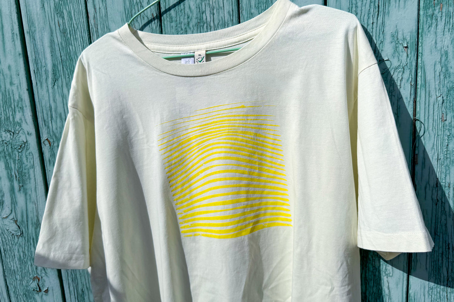 Detailansicht von einem hellgelben T-Shirt in Größe XL vor einem blauen Hintergrund. Das T-Shirt hat einen welligen Druck in gelb auf der Brust. Handbedruckt von Jeff - EveryPieceAFriend