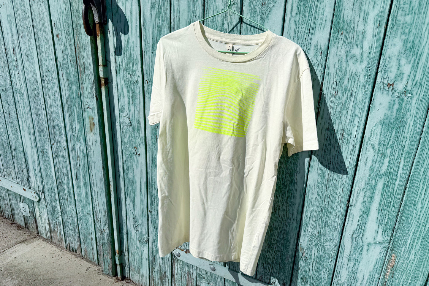 Detailansicht von einem hellgelben T-Shirt vor in der Größe S vor einem blau Hintergrund. Das T-Shirt hat einen welligen, neongelben Druck auf der Brust. Handbedruckt von Jeff - EveryPieceAFriend
