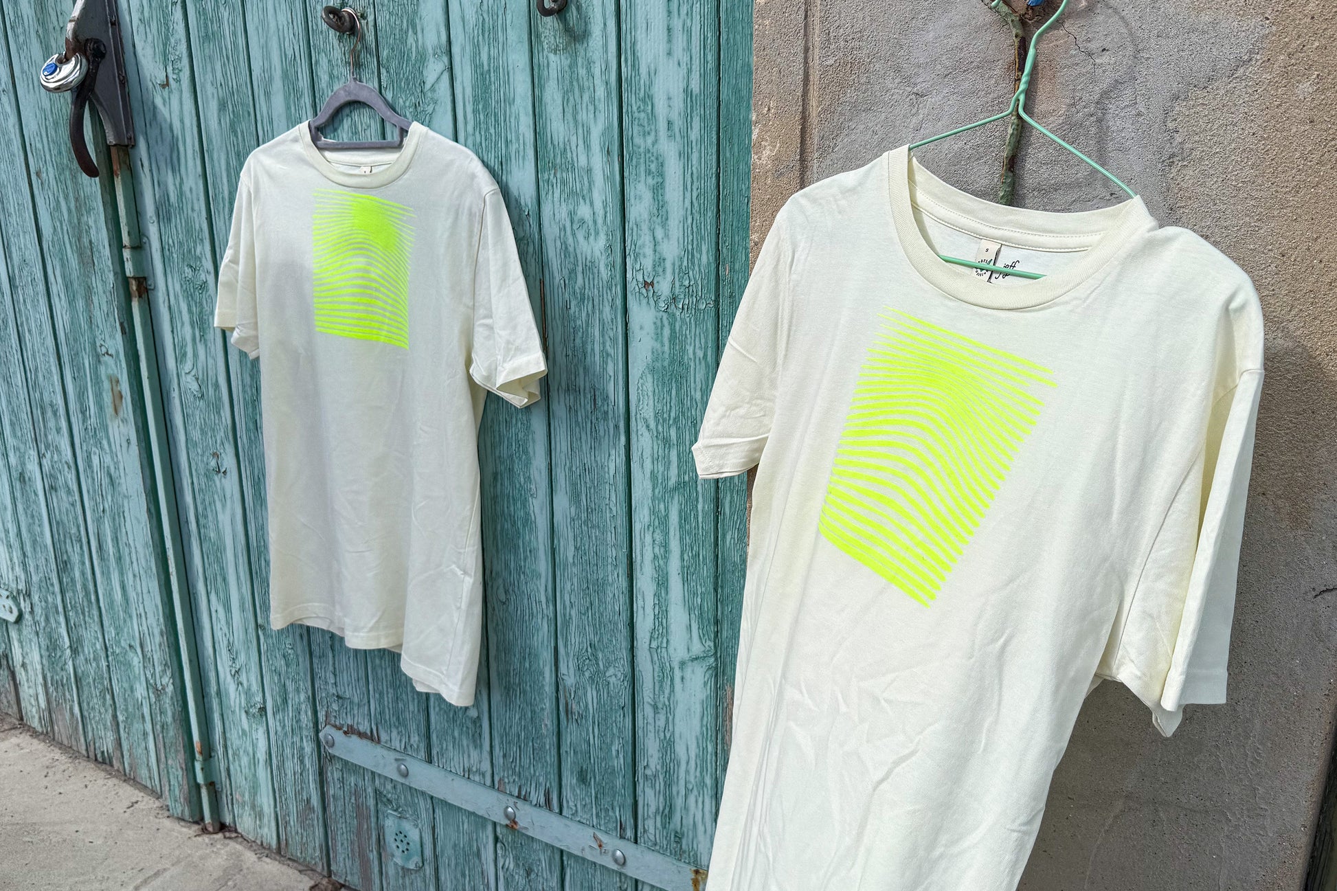 Detailansicht von zwei hellgelben T-Shirts in der Größe S vor einem blau-beigen Hintergrund. Die T-Shirts haben einen welligen, neongelben Druck auf der Brust. Handbedruckt von Jeff - EveryPieceAFriend