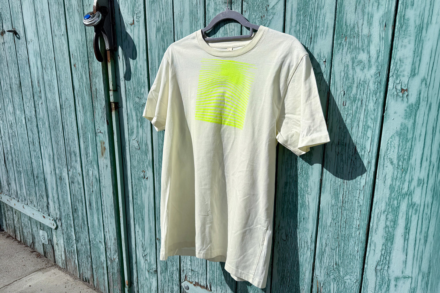 Detailansicht von einem hellgelben T-Shirt in der Größe S vor einem blauen Hintergrund. Das T-Shirt hat einen welligen, neongelben Druck auf der Brust. Handbedruckt von Jeff - EveryPieceAFriend