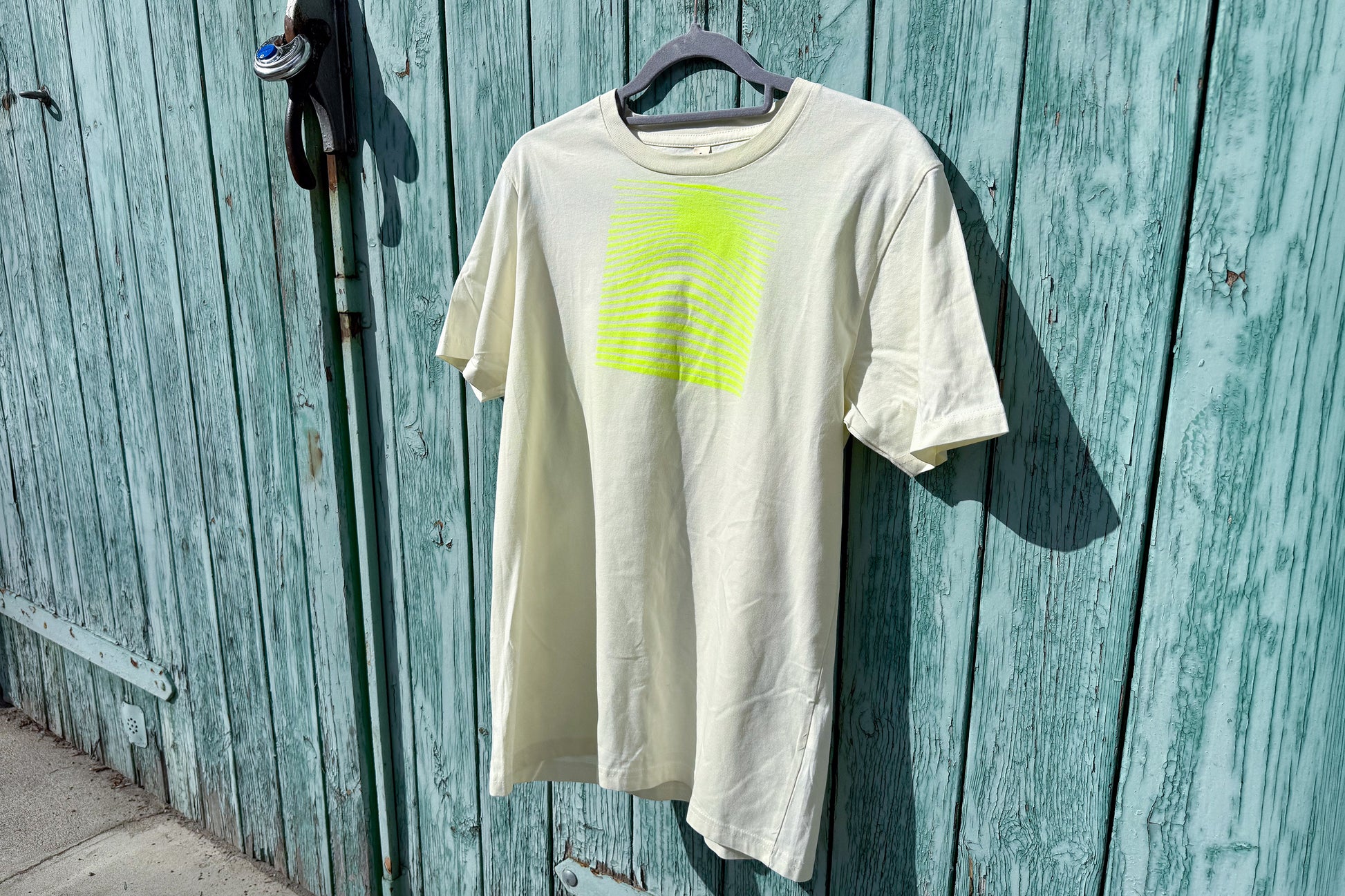 Detailansicht von einem hellgelben T-Shirt in der Größe S vor einem blauen Hintergrund. Das T-Shirt hat einen welligen, neongelben Druck auf der Brust. Handbedruckt von Jeff - EveryPieceAFriend