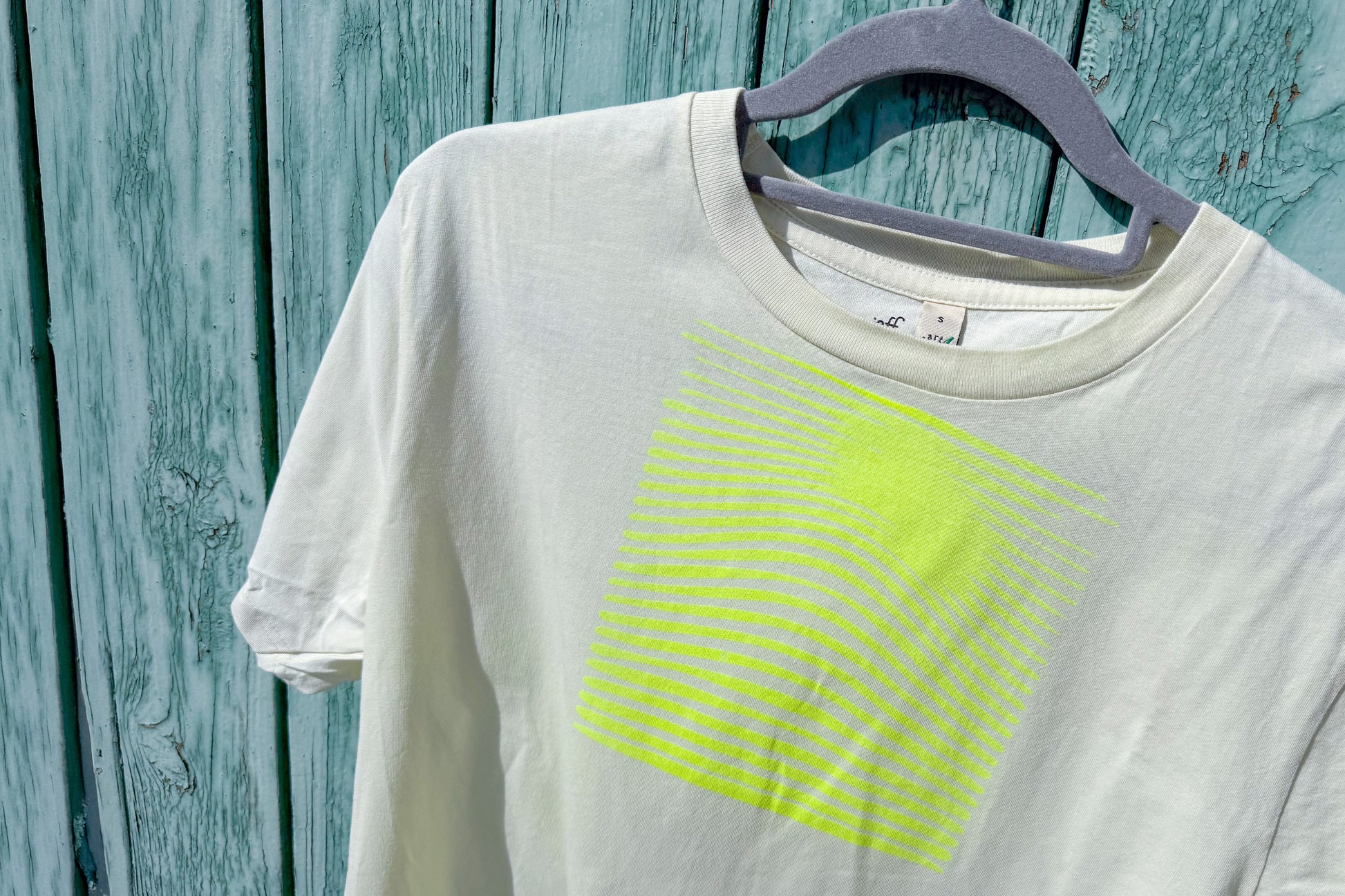 Detailansicht von einem hellgelben T-Shirt in der Größe S vor einem blauen Hintergrund. Das T-Shirt hat einen welligen, neongelben Druck auf der Brust. Handbedruckt von Jeff - EveryPieceAFriend