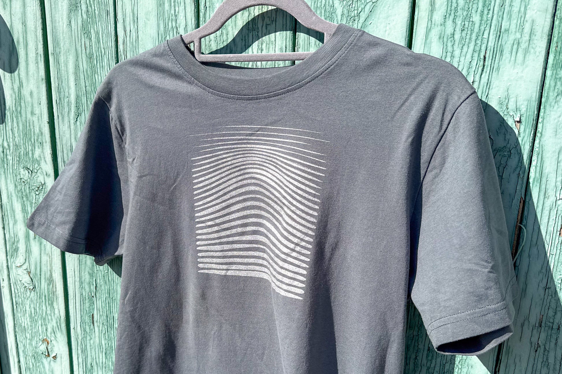Detailansicht von einem grauen T-Shirt in der Größe S vor einem blauen Hintergrund. Das T-Shirt hat einen welligen, silbernen Druck auf der Brust. Handbedruckt von Jeff - EveryPieceAFriend