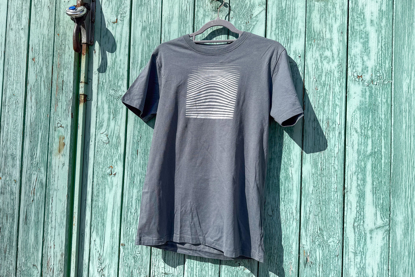 Ansicht von einem grauen T-Shirt in der Größe S vor einem blauen Hintergrund. Das T-Shirt hat einen welligen, silbernen Druck auf der Brust. Handbedruckt von Jeff - EveryPieceAFriend