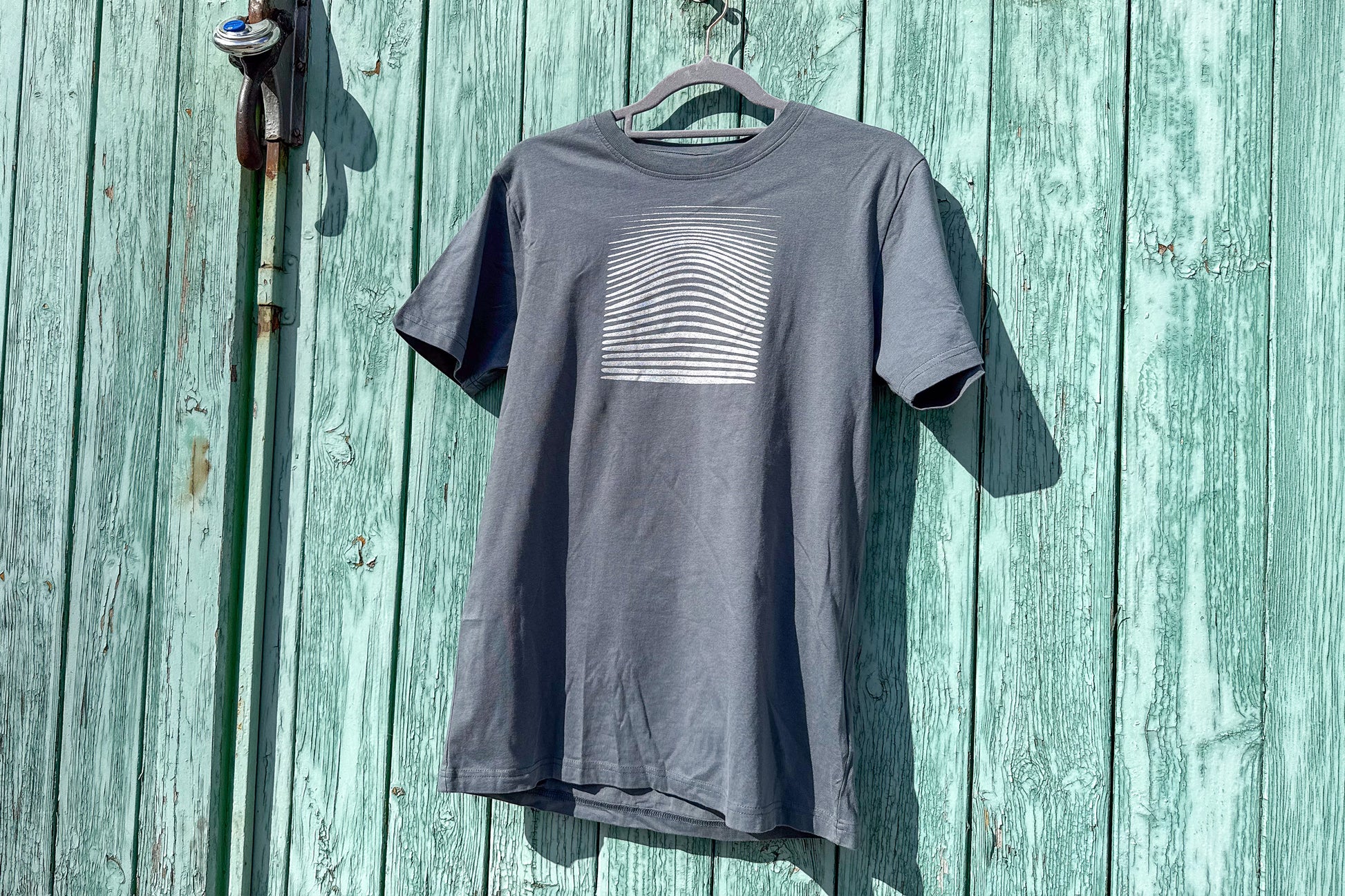 Ansicht von einem grauen T-Shirt in der Größe S vor einem blauen Hintergrund. Das T-Shirt hat einen welligen, silbernen Druck auf der Brust. Handbedruckt von Jeff - EveryPieceAFriend