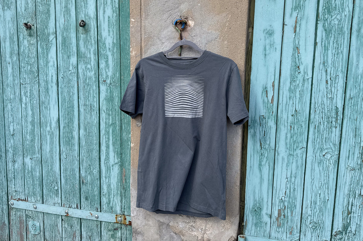 Detailansicht von einem grauen T-Shirt in der Größe S vor einem blau-beigen Hintergrund. Das T-Shirt hat einen welligen, silbernen Druck auf der Brust. Handbedruckt von Jeff - EveryPieceAFriend