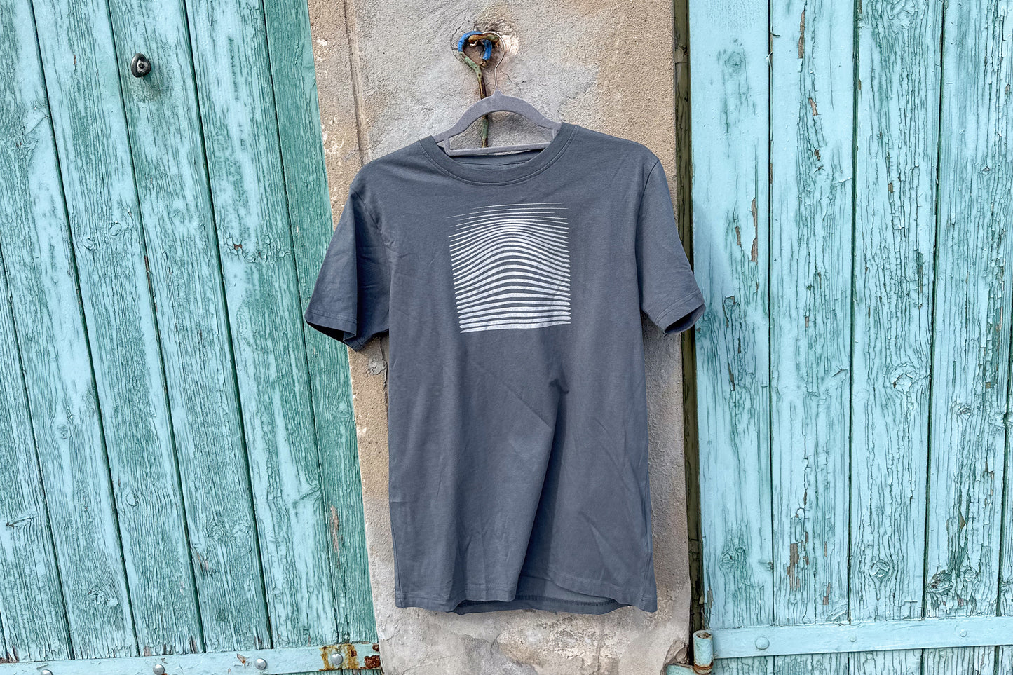 Ansicht von einem grauen T-Shirt in der Größe S vor einem blau-beigen Hintergrund. Das T-Shirt hat einen welligen, silbernen Druck auf der Brust. Handbedruckt von Jeff - EveryPieceAFriend