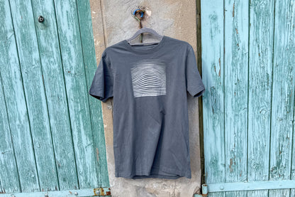 Ansicht von einem grauen T-Shirt in der Größe S vor einem blau-beigen Hintergrund. Das T-Shirt hat einen welligen, silbernen Druck auf der Brust. Handbedruckt von Jeff - EveryPieceAFriend