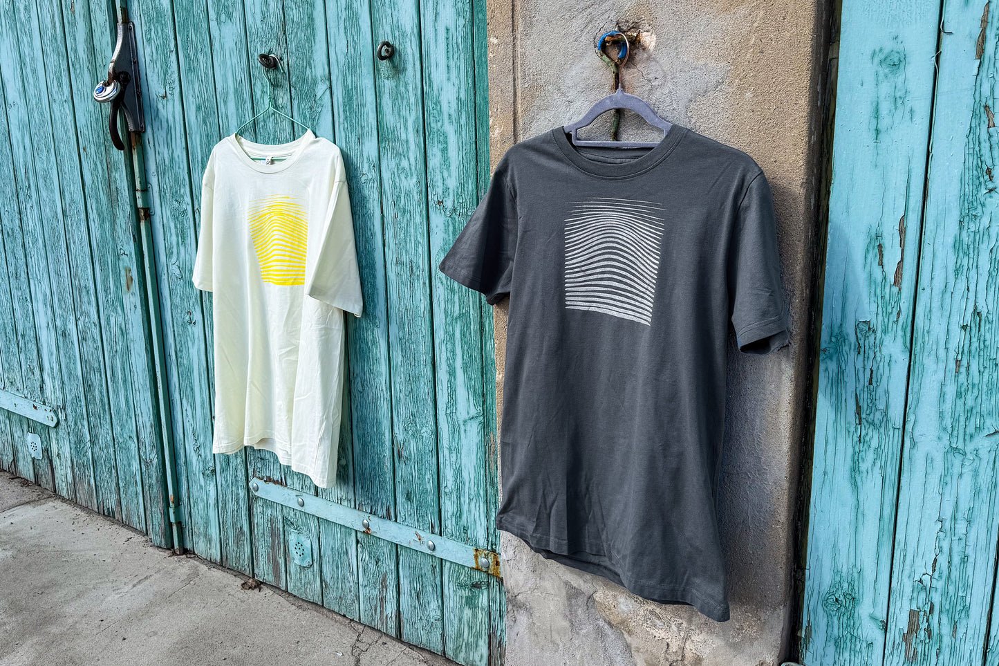 Ansicht von einem grauen und einem hellgelben T-Shirt vor einem blau-beigen Hintergrund. Das graue T-Shirt hat einen welligen Druck in silber, das hellgelbe Shirt einen welligen Druck in gelb auf der Brust. Handbedruckt von Jeff - EveryPieceAFriend