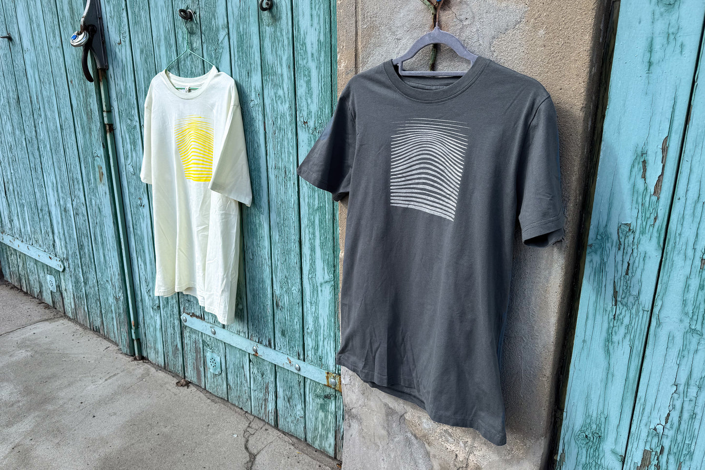 Ansicht von einem grauen und einem hellgelben T-Shirt vor einem blau-beigen Hintergrund. Das graue T-Shirt hat einen welligen Druck in silber, das hellgelbe Shirt einen welligen Druck in gelb auf der Brust. Handbedruckt von Jeff - EveryPieceAFriend