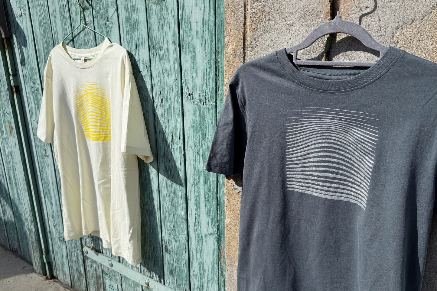 Ansicht von einem grauen und einem hellgelben T-Shirt vor einem blau-beigen Hintergrund. Das graue T-Shirt hat einen welligen Druck in silber, das hellgelbe Shirt einen welligen Druck in gelb auf der Brust. Handbedruckt von Jeff - EveryPieceAFriend