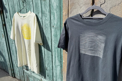 Ansicht von einem grauen und einem hellgelben T-Shirt vor einem blau-beigen Hintergrund. Das graue T-Shirt hat einen welligen Druck in silber, das hellgelbe Shirt einen welligen Druck in gelb auf der Brust. Handbedruckt von Jeff - EveryPieceAFriend
