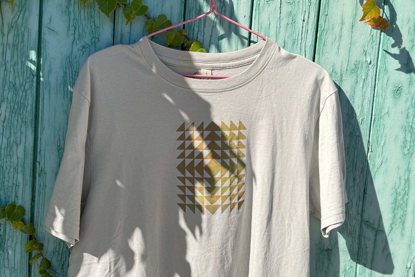 Detailansicht: Vorderseite eines T-Shirt in hellem grau vor blauen Hintergrund. Das Shirt zeigt ein minimalistisches und geometrisches Muster in der Farbe ocker auf der Brust.