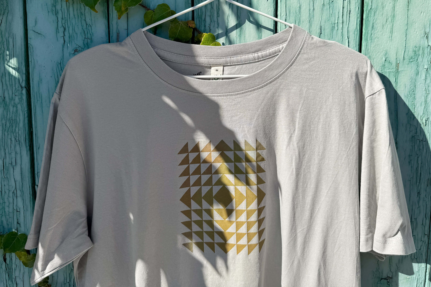 Detailansicht: Vorderseite eines T-Shirt in hellem grau vor blauen Hintergrund. Das Shirt zeigt ein minimalistisches und geometrisches Muster in der Farbe ocker auf der Brust.