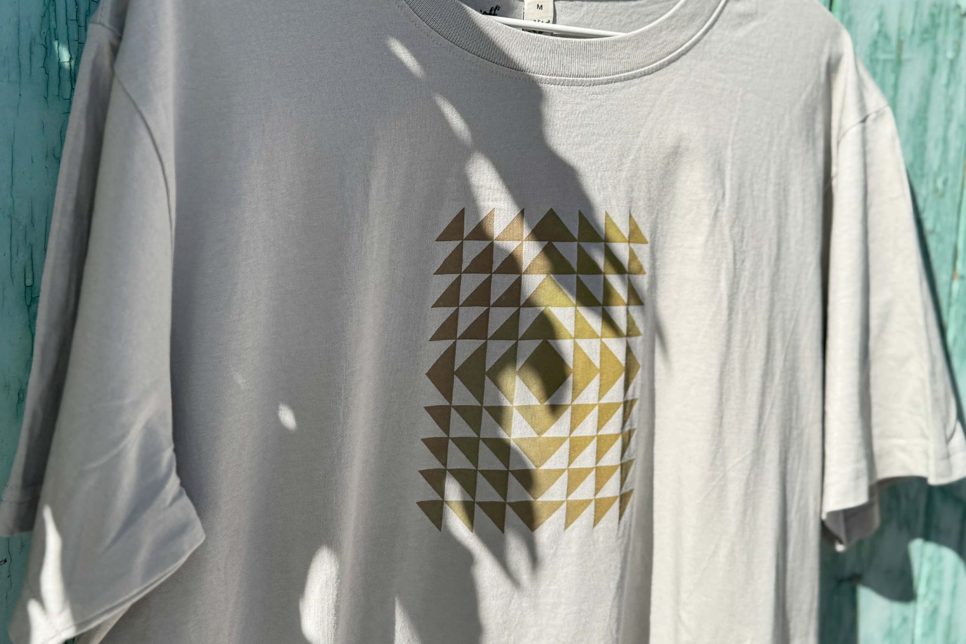 Detailansicht: Vorderseite eines T-Shirt in hellem grau vor blauen Hintergrund. Das Shirt zeigt ein minimalistisches und geometrisches Muster in der Farbe ocker auf der Brust.