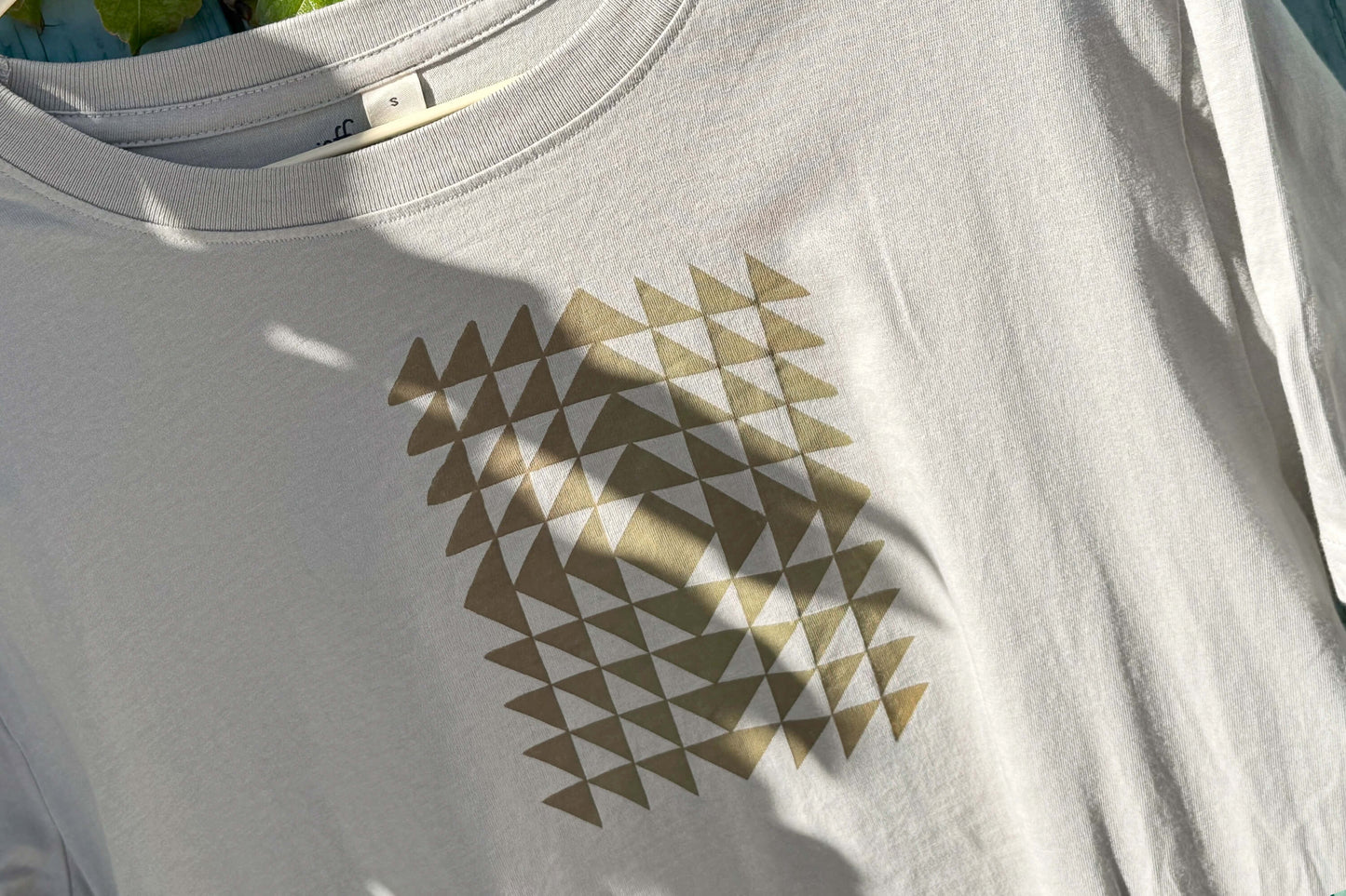 Detailansicht: Vorderseite eines T-Shirt in hellem grau vor blauen Hintergrund. Das Shirt zeigt ein minimalistisches und geometrisches Muster in der Farbe ocker auf der Brust.