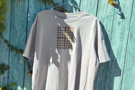 Detailansicht: Vorderseite eines T-Shirt in hellem grau vor blauen Hintergrund. Das Shirt zeigt ein minimalistisches und geometrisches Muster in der Farbe ocker auf der Brust.