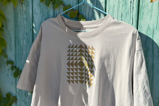 Detailansicht: Vorderseite eines T-Shirt in hellem grau vor blauen Hintergrund. Das Shirt zeigt ein minimalistisches und geometrisches Muster in der Farbe ocker auf der Brust.