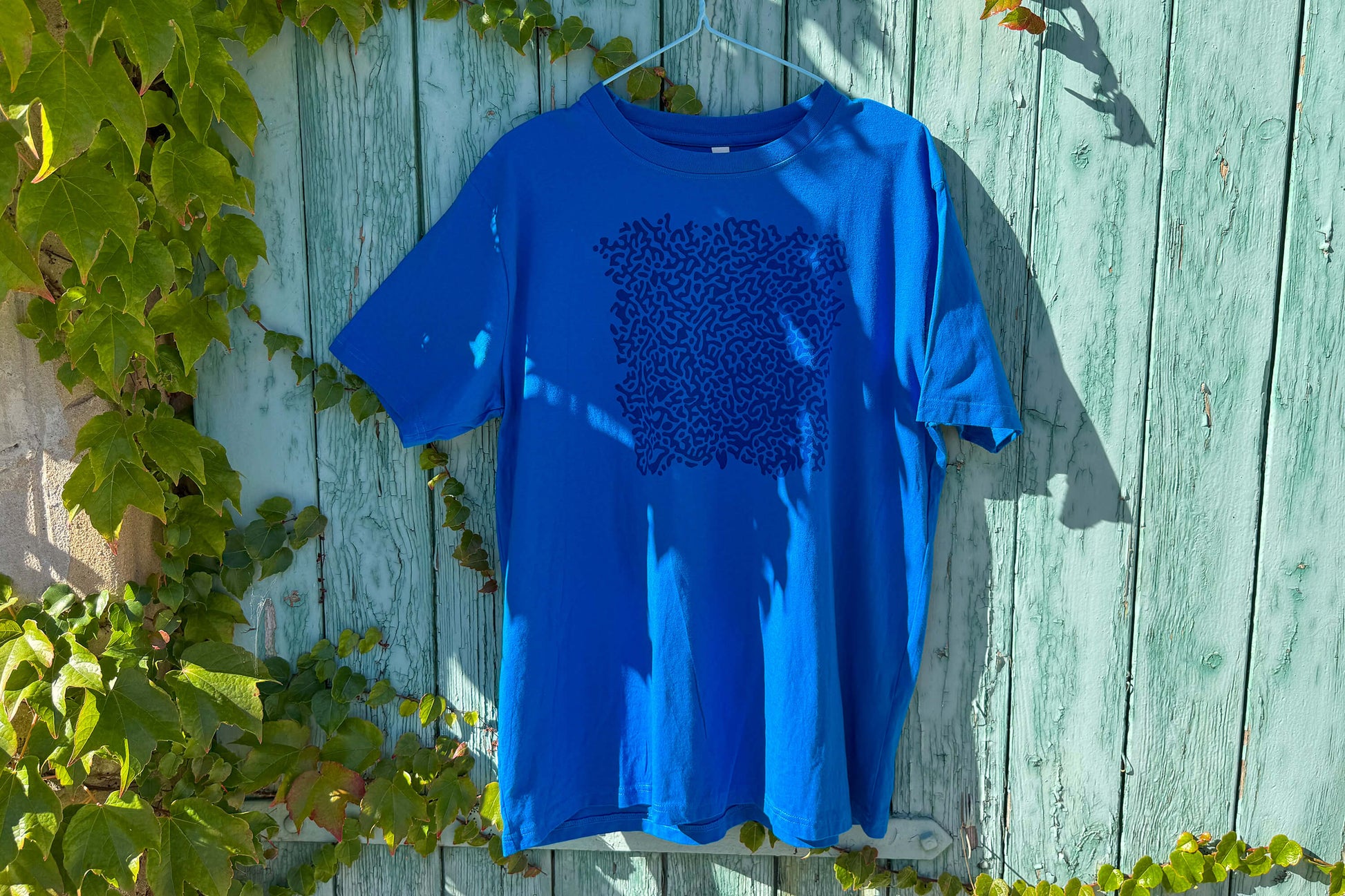 Gesamtansicht von einem blauen T-Shirt vor einem blauen Hintergrund. Das T-Shirt hat einen welligen Druck auf der Brust.