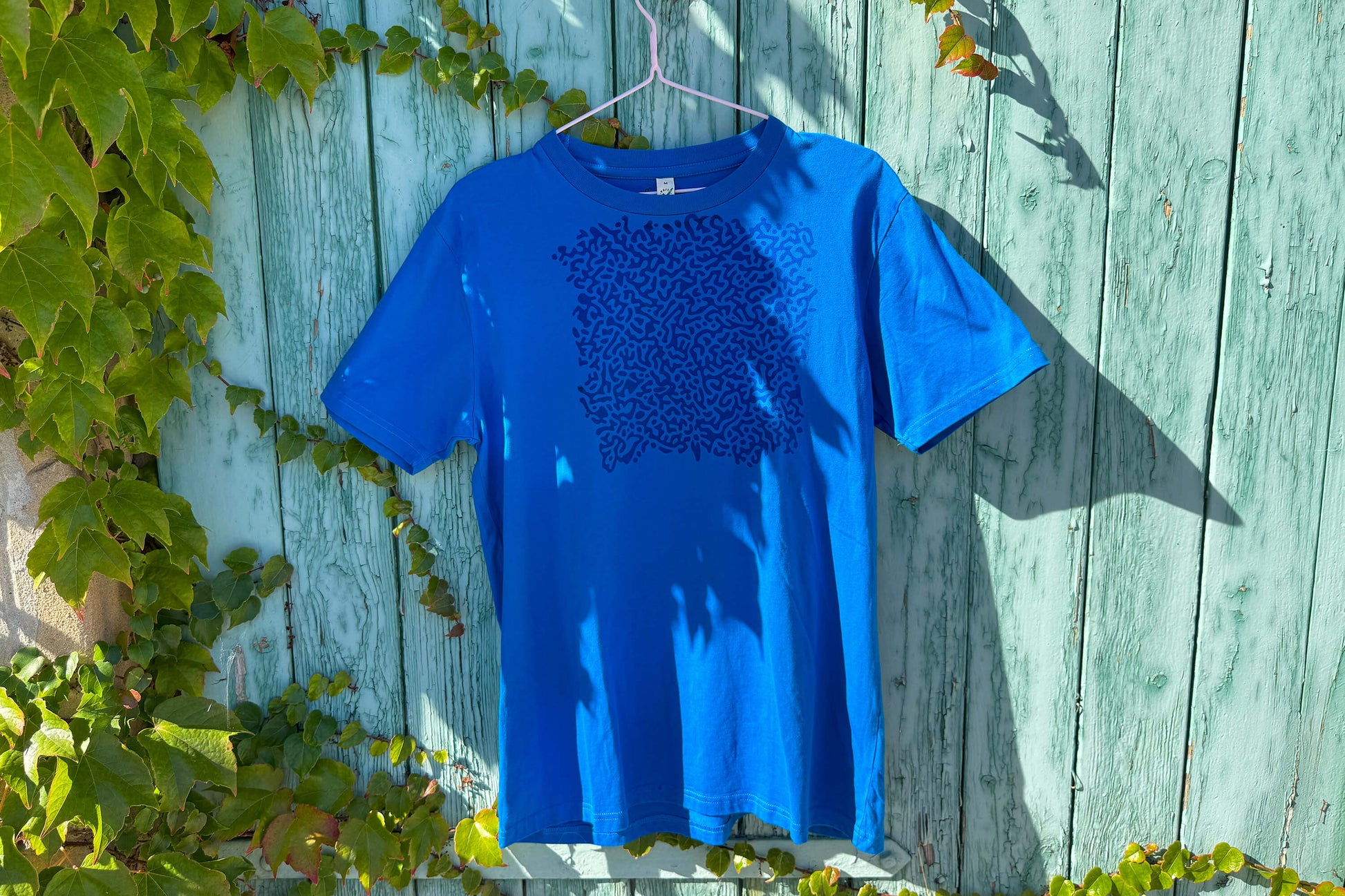 Gesamtansicht von einem blauen T-Shirt vor einem blauen Hintergrund. Das T-Shirt hat einen welligen Druck auf der Brust.