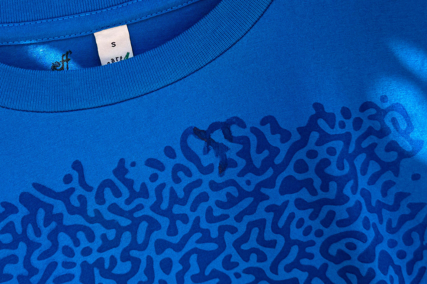 Detailansicht von einem blauen T-Shirt in der Größe S. Das T-Shirt hat einen welligen Druck auf der Brust.