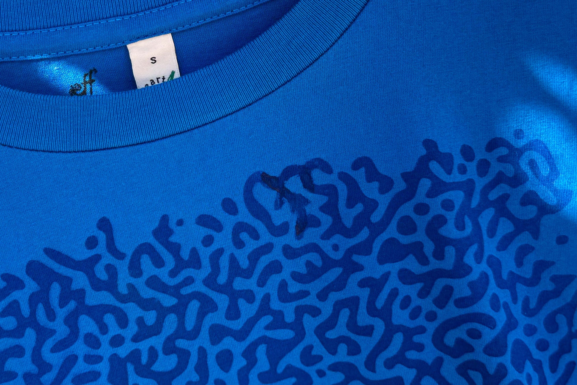 Detailansicht von einem blauen T-Shirt in der Größe S. Das T-Shirt hat einen welligen Druck auf der Brust.