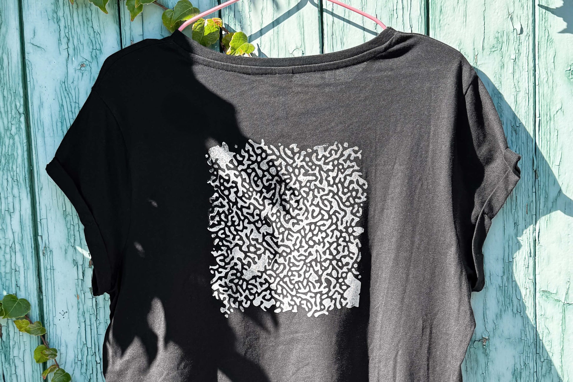 Hintere Detailansicht von einem schwarzen T-Shirt vor einem blauen Hintergrund. Das T-Shirt hat einen welligen Druck in silberner Farbe auf dem Rücken.