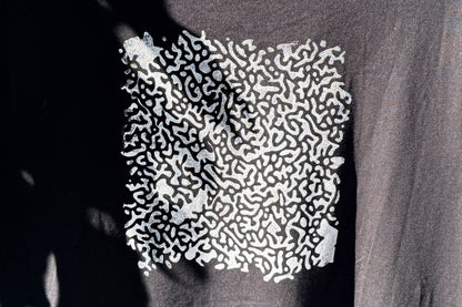 Hintere Detailansicht von einem schwarzen T-Shirt. Das T-Shirt hat einen welligen Druck in silberner Farbe auf dem Rücken.