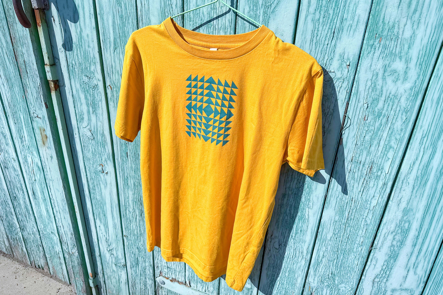 Gesamtansicht: Vorderseite eines T-Shirt in knalligem gelb vor blauen Hintergrund. Das Shirt zeigt ein minimalistisches und geometrisches Muster in der Farbe blau auf der Brust.