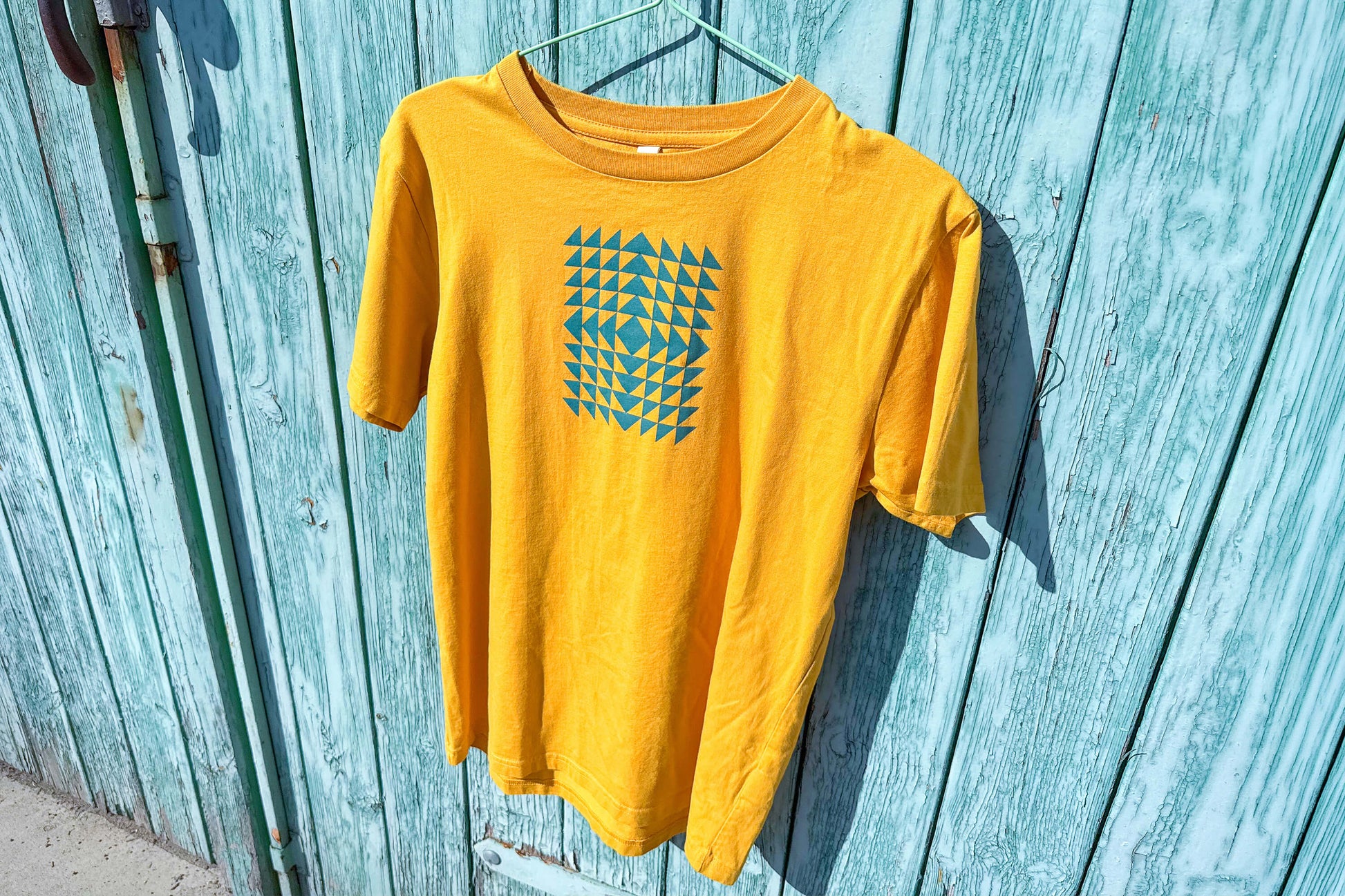 Gesamtansicht: Vorderseite eines T-Shirt in knalligem gelb vor blauen Hintergrund. Das Shirt zeigt ein minimalistisches und geometrisches Muster in der Farbe blau auf der Brust.