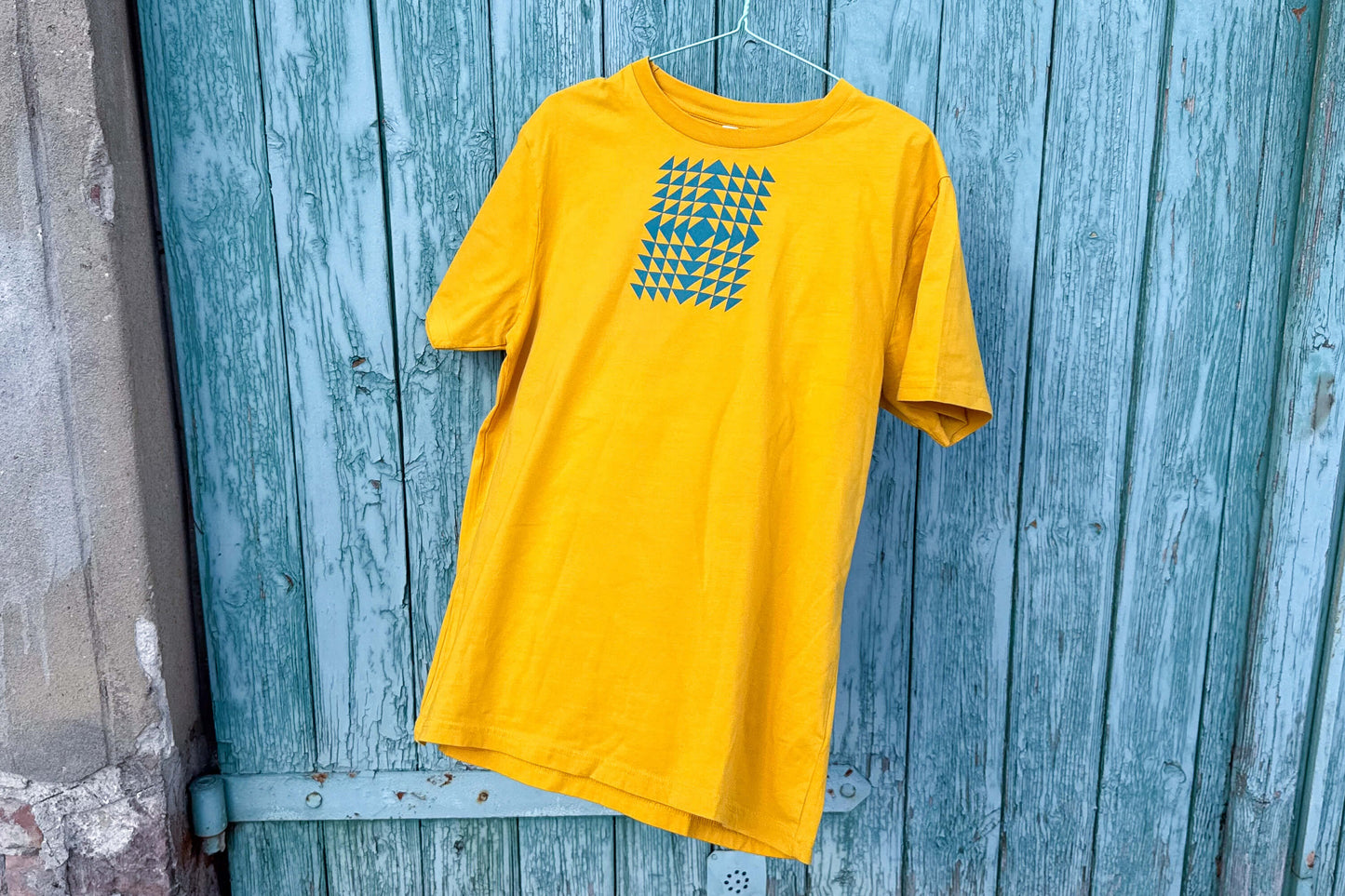 Gesamtansicht: Vorderseite eines T-Shirt in knalligem gelb vor blauen Hintergrund. Das Shirt zeigt ein minimalistisches und geometrisches Muster in der Farbe blau auf der Brust.