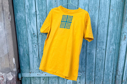 Gesamtansicht: Vorderseite eines T-Shirt in knalligem gelb vor blauen Hintergrund. Das Shirt zeigt ein minimalistisches und geometrisches Muster in der Farbe blau auf der Brust.