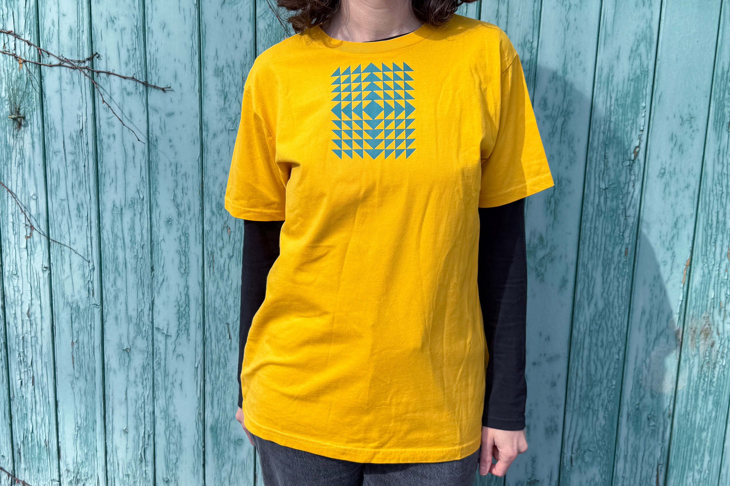 Gesamtansicht: Frau trägt ein T-Shirt in knalligem gelb vor blauen Hintergrund. Das Shirt zeigt ein minimalistisches und geometrisches Muster in der Farbe blau auf der Brust.
