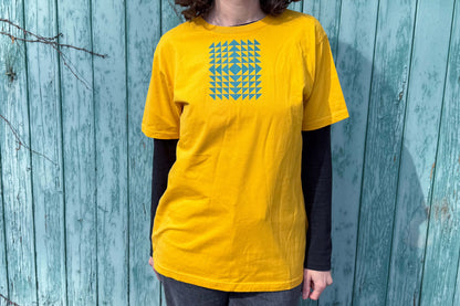 Gesamtansicht: Frau trägt ein T-Shirt in knalligem gelb vor blauen Hintergrund. Das Shirt zeigt ein minimalistisches und geometrisches Muster in der Farbe blau auf der Brust.