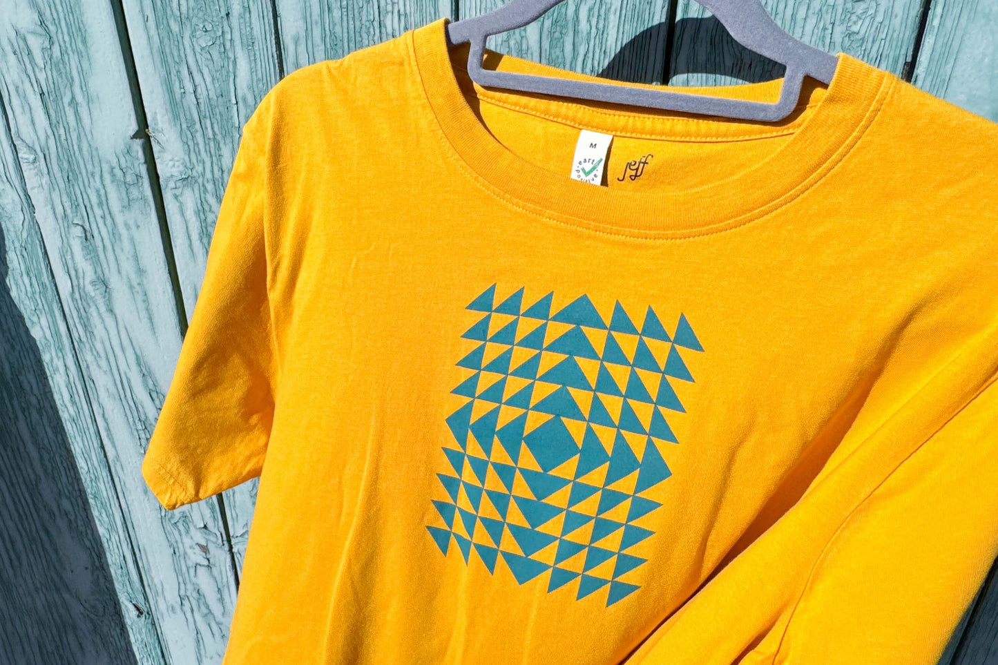 Detailansicht: Vorderseite eines T-Shirt in knalligem gelb vor blauen Hintergrund. Das Shirt zeigt ein minimalistisches und geometrisches Muster in der Farbe blau auf der Brust.