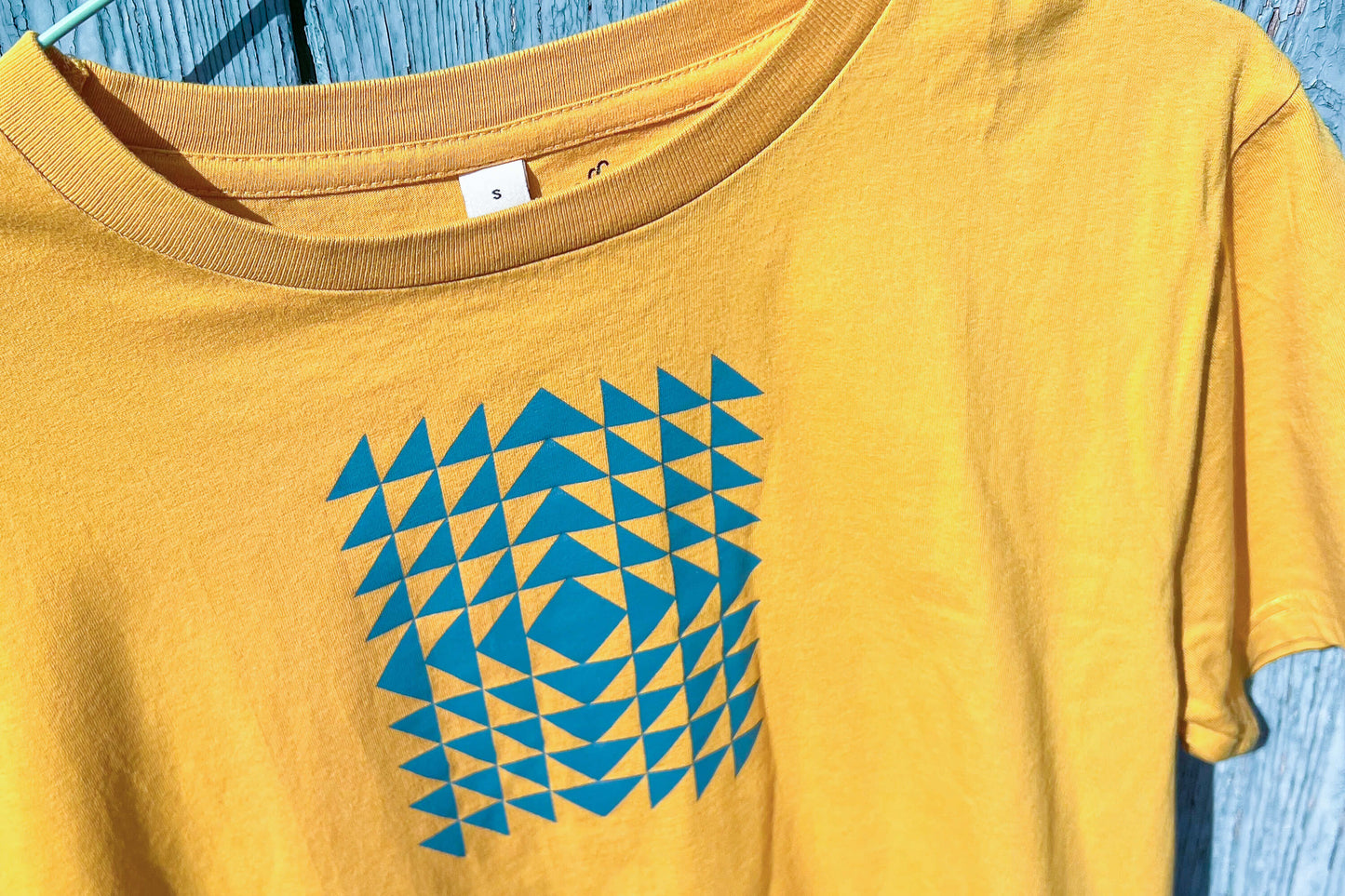 Detailansicht: Vorderseite eines T-Shirt in knalligem gelb vor blauen Hintergrund. Das Shirt zeigt ein minimalistisches und geometrisches Muster in der Farbe blau auf der Brust.