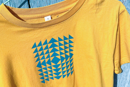 Detailansicht: Vorderseite eines T-Shirt in knalligem gelb vor blauen Hintergrund. Das Shirt zeigt ein minimalistisches und geometrisches Muster in der Farbe blau auf der Brust.