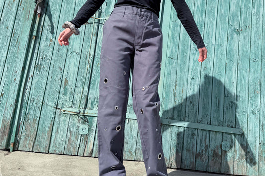 Vorderansicht einer Frau, sie trägt eine graue Hose mit silbernen Ösen und Nieten. Sie steht vor einem hellblauer Garagentor. jeff-everyPieceAFriend