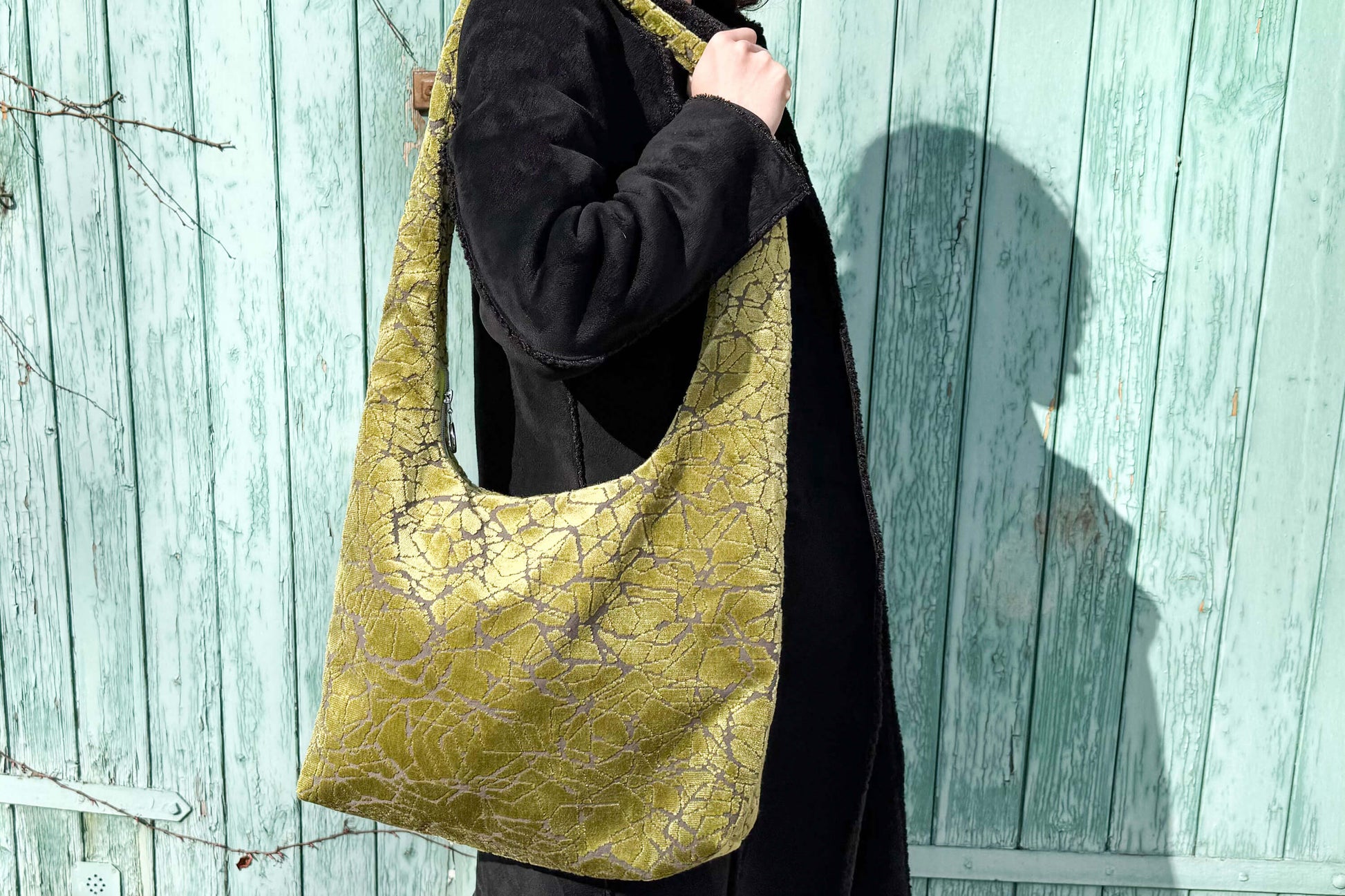 Große Crossbody Tasche in grünem Muster und silbernen Reißverschluss steht vor hellblauem Holz-Hintergrund. 