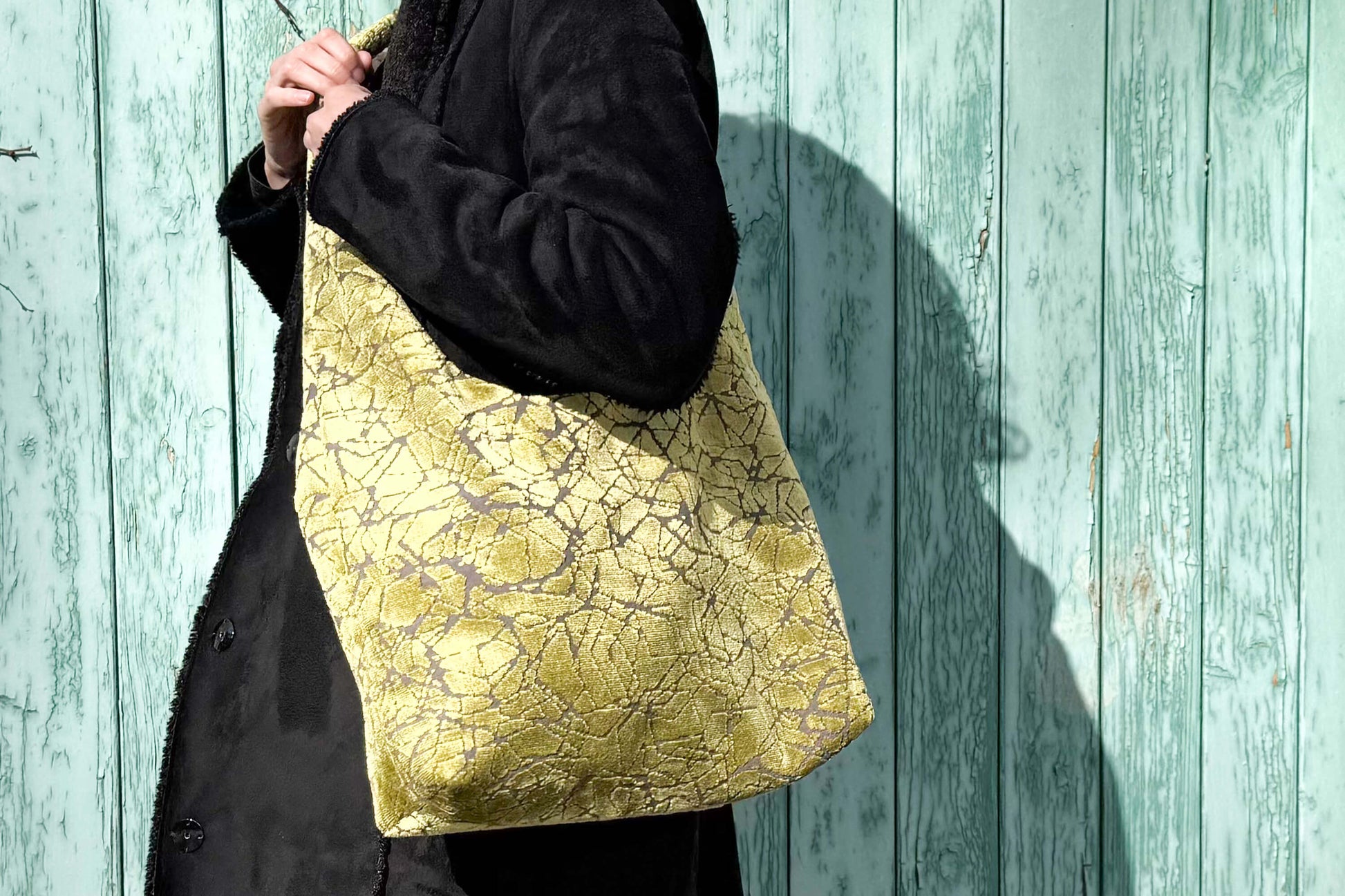 Große Crossbody Tasche in grünem Muster und silbernen Reißverschluss steht vor hellblauem Holz-Hintergrund
