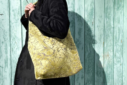 Große Crossbody Tasche in grünem Muster und silbernen Reißverschluss steht vor hellblauem Holz-Hintergrund