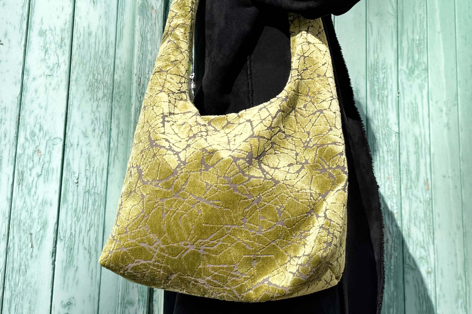 Zoomansicht große Crossbody Tasche in grünem Muster und silbernen Reißverschluss steht vor hellblauem Holz-Hintergrund. 