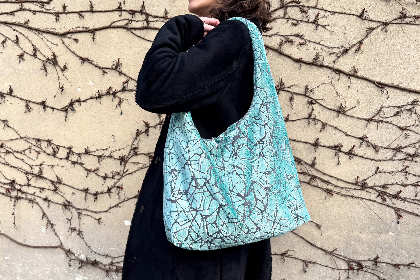 Eine Frau mit schwarzen Mantel und einer hellblauen, großen Hobo Bag, steht seitlich vor einer Wand. 