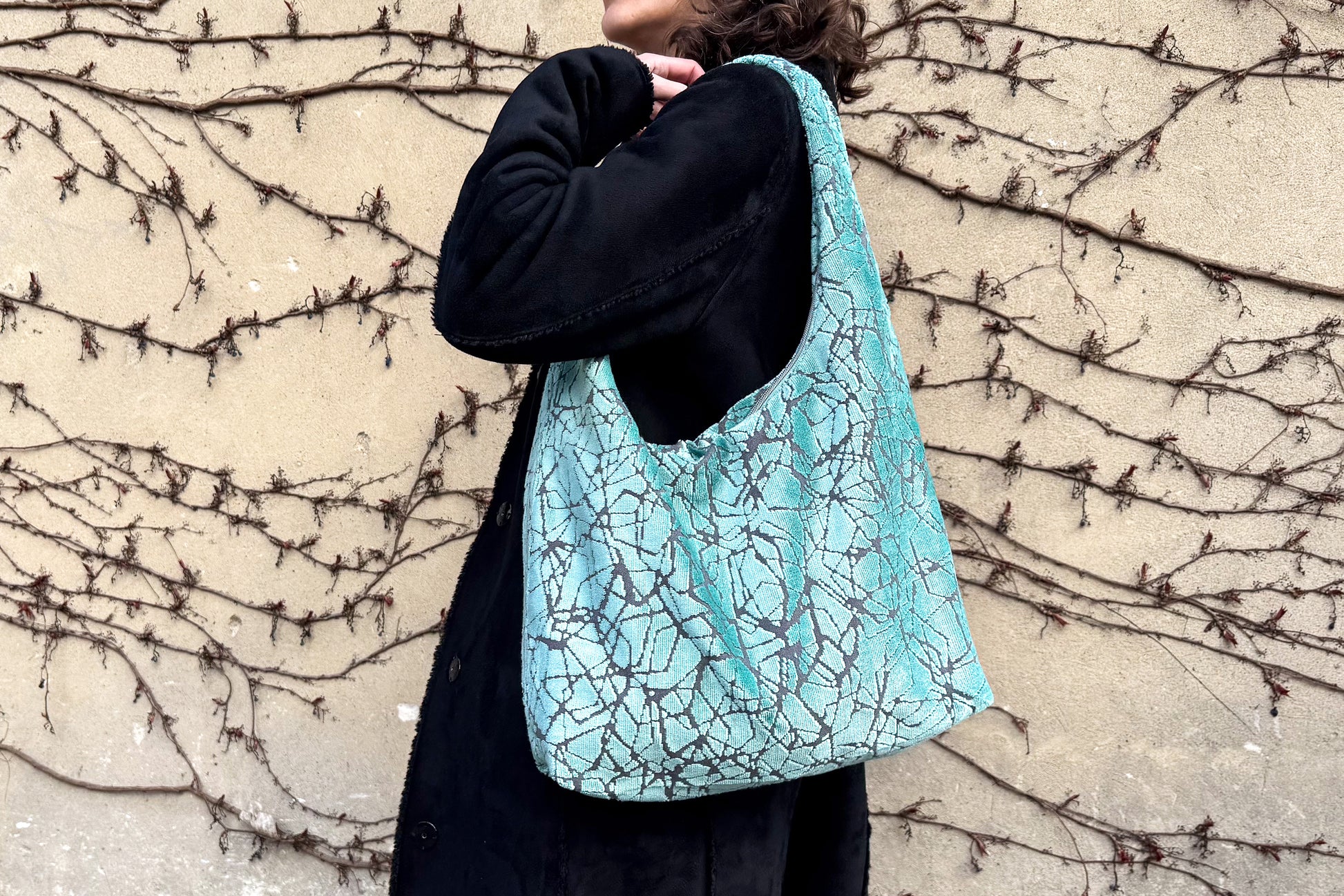Eine Frau mit schwarzen Mantel und einer hellblauen, großen Hobo Bag, steht seitlich vor einer Wand. 