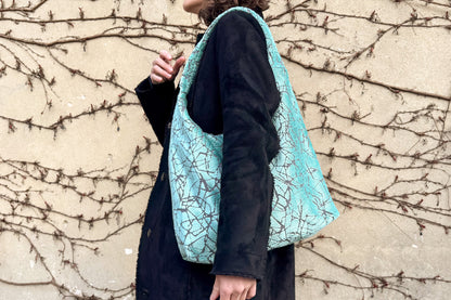 Eine Frau mit schwarzen Mantel und einer hellblauen, großen Hobo Bag, steht seitlich vor einer Wand. 